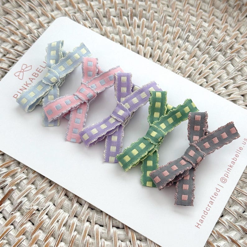 May include: A set of six pastel-colored hair bows with a checkered pattern. The bows are in shades of blue, pink, purple, green, and gray, and are arranged on a white card. The card has the text "PINKABELLE" and "Handcrafted | @pinkabelle.us".