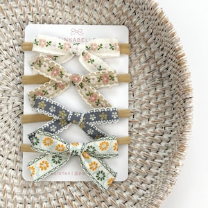 May include: A set of four baby headbands with floral bow accents. The bows feature various patterns, including pink and white flowers, and orange and green floral designs. The headbands are displayed on a white card with the brand name "PINKABELI".