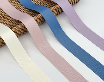 May include: Close-up of several solid-colored ribbons in shades of cream, mauve, blue, and lavender. The ribbons are arranged diagonally, with a woven basket in the background. The ribbons have a ribbed texture.