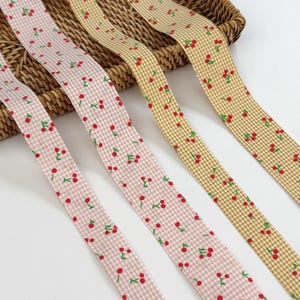 May include: Four ribbons with a gingham pattern and cherry print. The ribbons are in shades of pink and tan. The cherries are red with green leaves. The ribbons are arranged diagonally across the image.