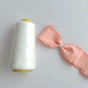 May include: A white thread spool with a yellow base and top, next to a light pink fabric bow with ruffled edges. The items are on a white background. The thread spool is wrapped in clear plastic.