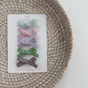 May include: A set of six colorful gingham hair bows in pastel shades of blue, pink, purple, green, and gray. The bows are attached to a white card with the text "PINKABELLE" and "Handcrafted | @pinkabelle.us".