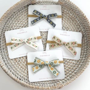 May include: Five floral fabric bows on headbands, displayed on white cards inside a woven basket. The bows feature various colors, including blue, cream, and yellow, with delicate floral patterns and the brand name "PINKABELLE".