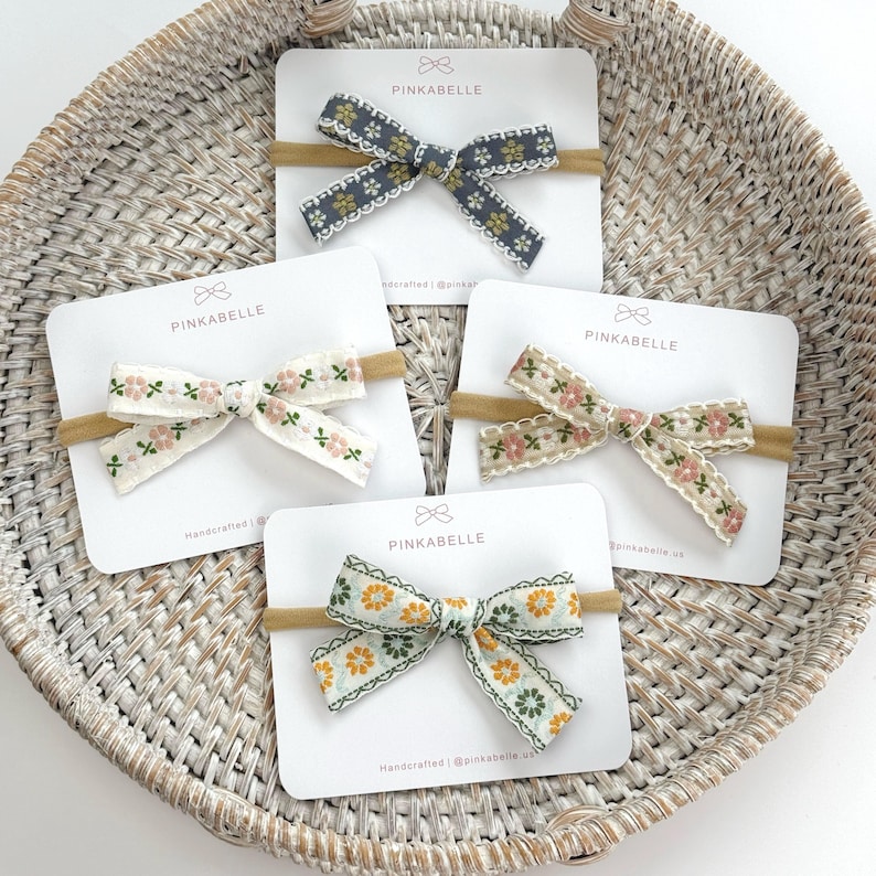 May include: Five floral fabric bows on headbands, displayed on white cards inside a woven basket. The bows feature various colors, including blue, cream, and yellow, with delicate floral patterns and the brand name "PINKABELLE".