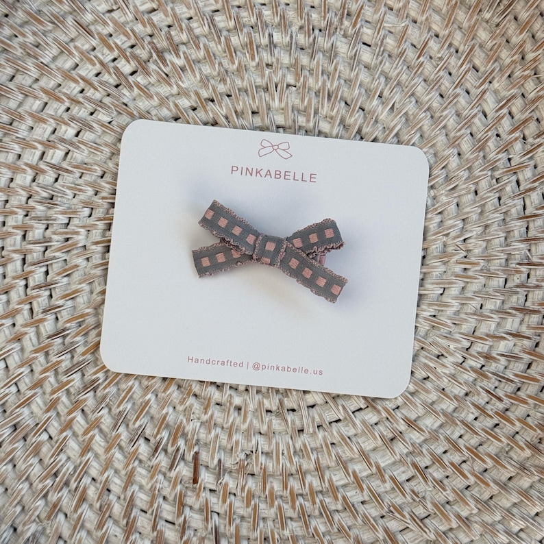 May include: A gray and pink checkered bow hair clip on a white card with the brand name "PINKABELLE". The card is on a woven surface. The text "Handcrafted | @pinkabelle.us" is printed on the card.