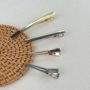 May include: Four metal hair clips in varying finishes including gold, silver, rose gold, and a matte silver tone. Each clip features a flat, elongated design with a hinged mechanism, arranged in a vertical stack on a textured surface.