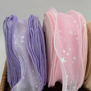 May include: Close-up of spools of decorative ribbon in pastel colors. The ribbons are purple and pink, with a sheer overlay featuring a pattern of small, shimmering stars. The edges of the ribbons are finished with a matching thread.