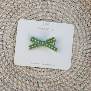May include: A green and yellow plaid bow hair clip on a white card. The card has the text "PINKABELLE" and "Handcrafted | @pinkabelle.us". The bow is made of a ribbon with a checkered pattern.