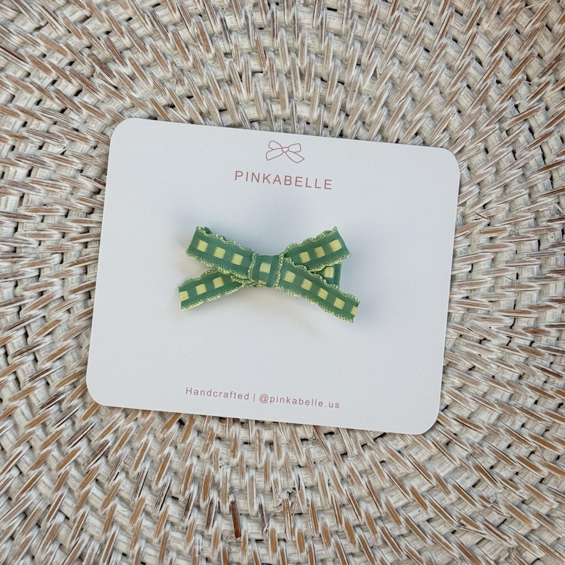 May include: A green and yellow plaid bow hair clip on a white card. The card has the text "PINKABELLE" and "Handcrafted | @pinkabelle.us". The bow is made of a ribbon with a checkered pattern.