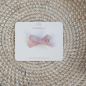 May include: A pink and gray bow hair clip with a heart pattern, presented on a white card. The card features the brand name "PINKABELLE" and the text "Handcrafted | @pinkabelle.us". The bow is made of a textured ribbon.