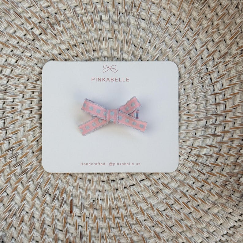 May include: A pink and gray bow hair clip with a heart pattern, presented on a white card. The card features the brand name "PINKABELLE" and the text "Handcrafted | @pinkabelle.us". The bow is made of a textured ribbon.