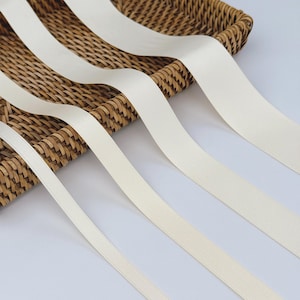 May include: Cream-coloured satin ribbons spilling from a woven basket. The ribbons have a smooth texture and a subtle sheen, giving a delicate and elegant look. These ribbons are suitable for crafts, gift wrapping, or adding a touch of sophistication to any project.