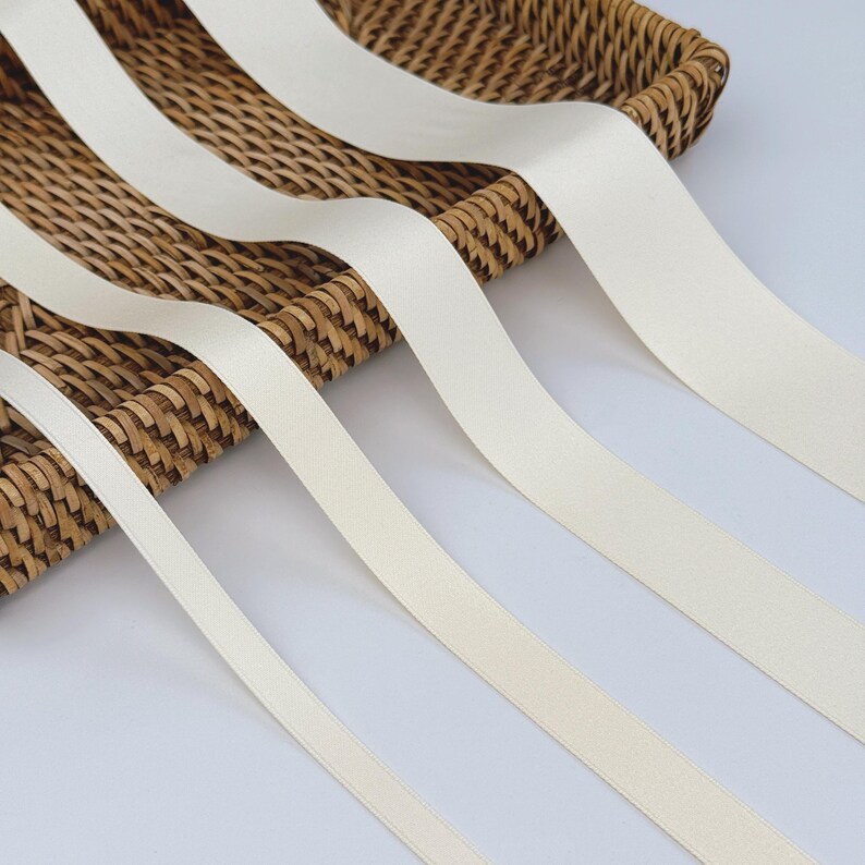 May include: Cream-coloured satin ribbons spilling from a woven basket. The ribbons have a smooth texture and a subtle sheen, giving a delicate and elegant look. These ribbons are suitable for crafts, gift wrapping, or adding a touch of sophistication to any project.