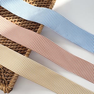 Könnte beinhalten: Drei Bänder mit Vichy-Muster in Hellblau, Pfirsich und Beige. Die Bänder sind diagonal über einem Weidenkorb platziert und bilden eine ansprechende Anordnung. Der Stoff hat ein klassisches Karomuster.