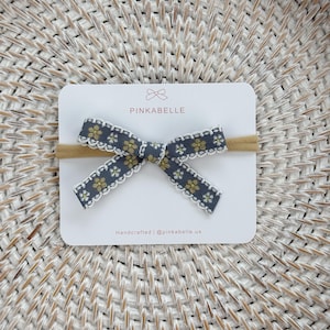 May include: A gray and yellow floral bow headband on a white card. The bow has a scalloped white edge and is attached to a tan elastic band. The card reads "PINKABELLE" and "Handcrafted | @pinkabelle.us".