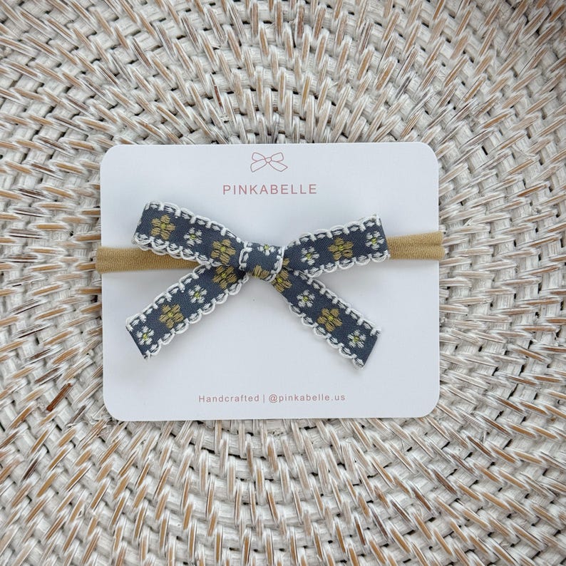 May include: A gray and yellow floral bow headband on a white card. The bow has a scalloped white edge and is attached to a tan elastic band. The card reads "PINKABELLE" and "Handcrafted | @pinkabelle.us".