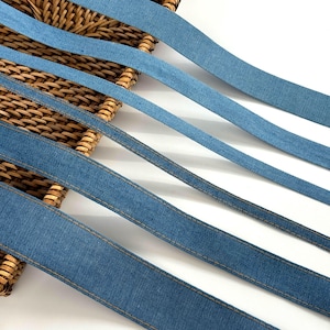 Op de afbeelding: Close-up van verschillende denim linten in verschillende blauwtinten. Elk lint is genaaid met een contrasterend gouden draad langs de randen. De linten zijn diagonaal gerangschikt tegen een geweven mandachtergrond, waardoor een textuur en gelaagd effect ontstaat.