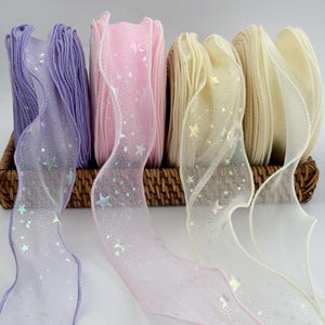 May include: A selection of decorative ribbons in a woven basket. The ribbons are in shades of purple, pink, and cream, with a sheer overlay adorned with silver star patterns. The spools of ribbon are tightly wound, showcasing their colors and textures.
