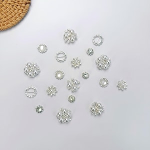 May include: A selection of decorative embellishments, featuring pearl and rhinestone floral and circular designs. The embellishments are in various sizes and shapes, with a bright, reflective appearance. The items are scattered on a white surface.