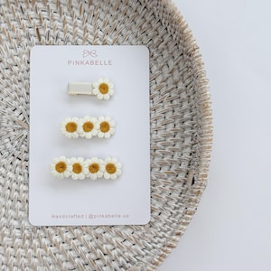 May include: A set of three daisy hair clips on a white card. The clips feature white petals and yellow centers. The card has the brand name "PINKABELLE" and the text "Handcrafted | @pinkabelle.us". The background is a woven basket.