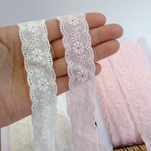 May include: Close-up of white and pink floral embroidered lace ribbons. The ribbons have a scalloped edge and are made of a delicate fabric. The white ribbon is held next to a pink ribbon, with a roll of pink lace in the background.