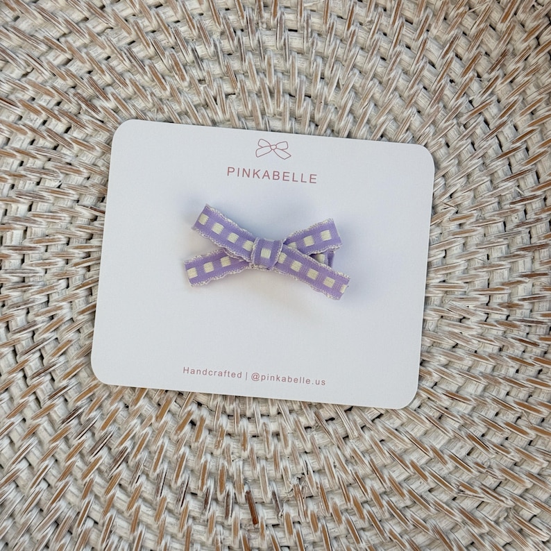 May include: A small, lavender and white checkered bow hair clip on a white card with the text "PINKABELLE". The card also includes the text "Handcrafted | @pinkabelle.us". The bow is made of fabric and has a decorative edge.