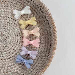 May include: A set of six small hair bows made of ribbed fabric, arranged in a vertical line. The bows feature a variety of soft pastel colors including white, pale yellow, peach, light pink, lavender, and light blue, each with a central knot.