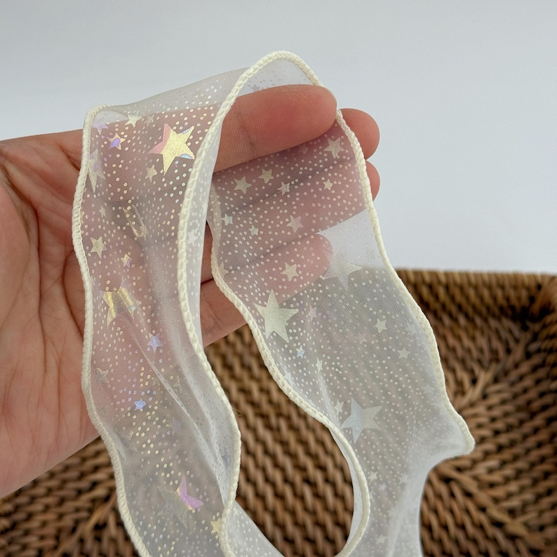May include: Sheer white ribbon with a cream-colored trim and a pattern of small white dots and iridescent stars. The ribbon is slightly wavy and appears to be made of a delicate material, suitable for crafts or gift wrapping.