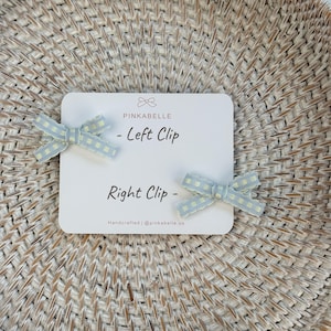 May include: Two light blue gingham bow hair clips, labeled "Left Clip" and "Right Clip" on a white card. The card also features the brand name "PINKABELLE" and the text "Handcrafted | @pinkabelle.us".