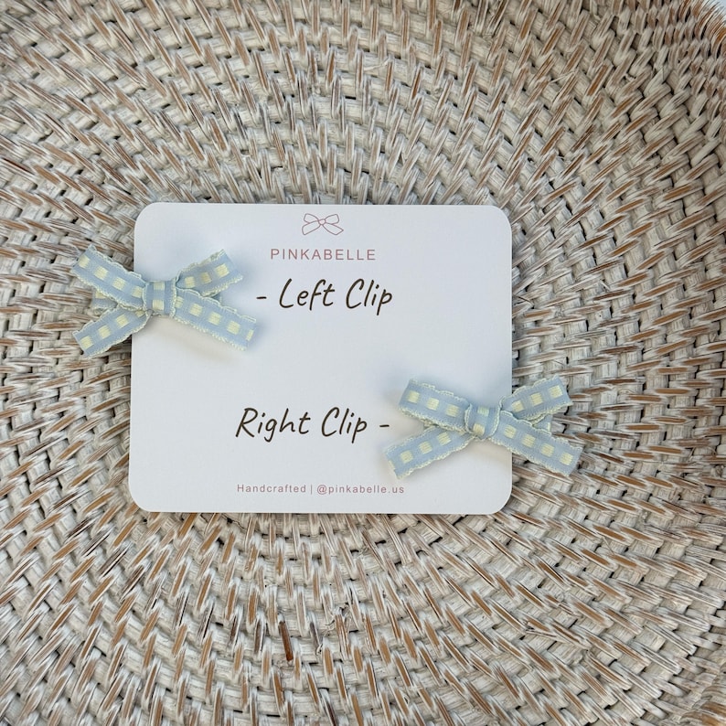 May include: Two light blue gingham bow hair clips, labeled "Left Clip" and "Right Clip" on a white card. The card also features the brand name "PINKABELLE" and the text "Handcrafted | @pinkabelle.us".