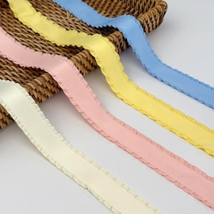 May include: Close-up of colorful ribbons in pastel shades of cream, pink, yellow, and blue. Each ribbon has a ruffled edge and a stitched border. The ribbons are arranged diagonally on a woven basket.