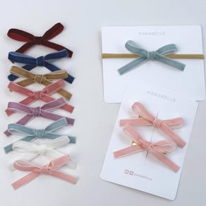May include: A selection of velvet hair bows in shades like burgundy, navy, tan, pink, lavender, and teal. Some are on clips, others on headbands. The packaging displays "PINKABELLE". Measurements are not applicable.
