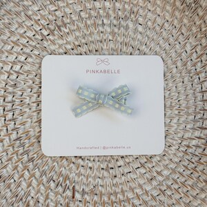 May include: A light blue and yellow gingham bow hair clip on a white card with the text "PINKABELLE". The card also includes the text "Handcrafted | @pinkabelle.us". The bow is a decorative accessory.