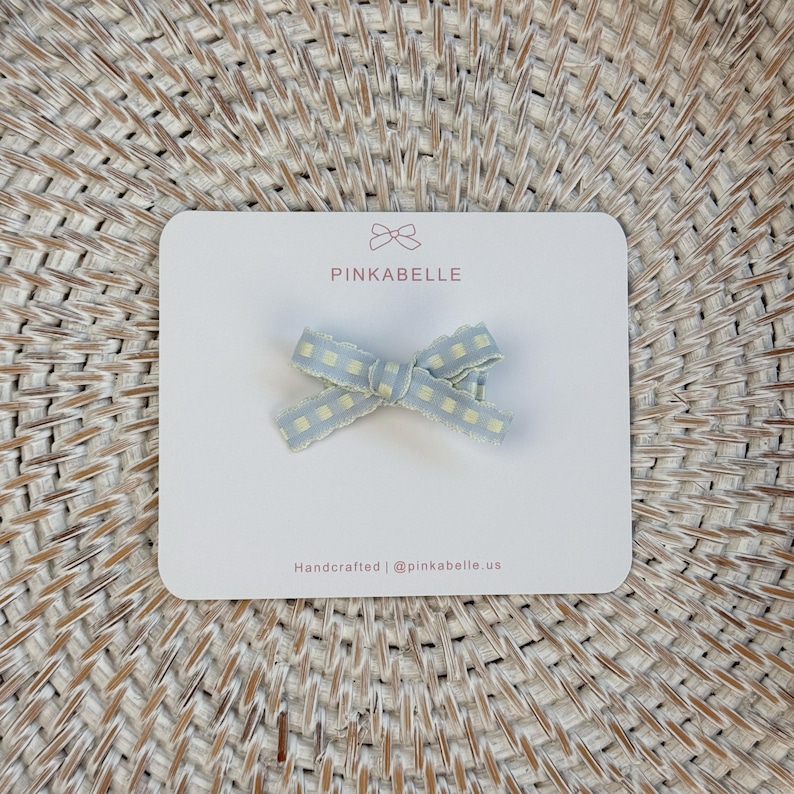 May include: A light blue and yellow gingham bow hair clip on a white card with the text "PINKABELLE". The card also includes the text "Handcrafted | @pinkabelle.us". The bow is a decorative accessory.
