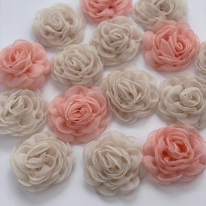 May include: A collection of fabric flowers in shades of blush pink and cream. The flowers are arranged in a scattered pattern, with each flower having multiple layers of petals. The petals have a soft, delicate texture.