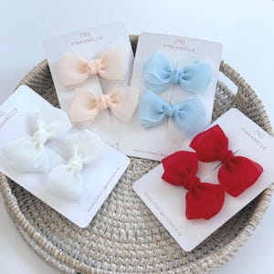 May include: A collection of hair bows in various colors, including white, peach, light blue, and red. Each bow is attached to a white card with the brand name "PINKABELLE" printed on it. The bows are arranged in a woven basket.