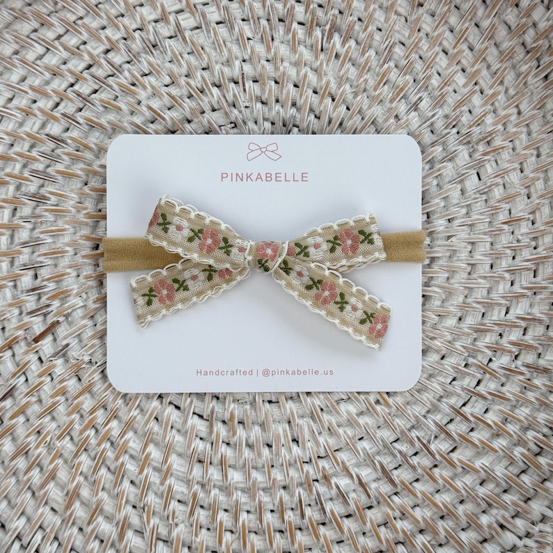 May include: A beige baby headband with a floral bow. The bow features a beige ribbon with pink flowers and green leaves, edged with white lace. The headband is on a white card with the text "PINKABELLE" and "Handcrafted | @pinkabelle.us".