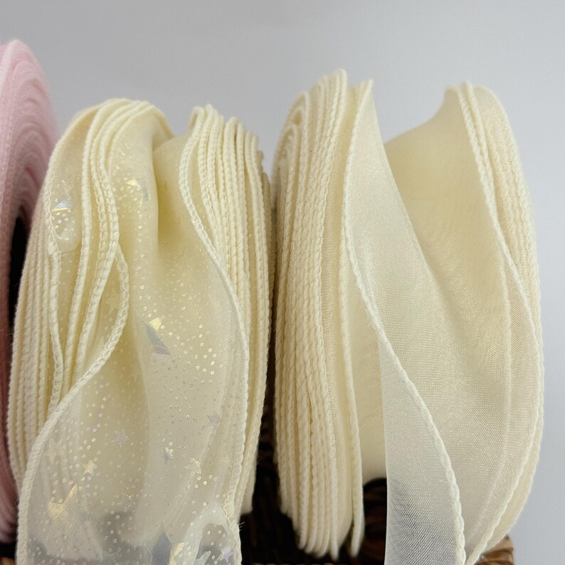 May include: Close-up of several rolls of decorative ribbon in shades of cream and pale pink. The cream ribbon has a sheer, sparkly overlay. The edges of the ribbon are stitched. The ribbons are likely used for crafts, gift wrapping, or decorations.