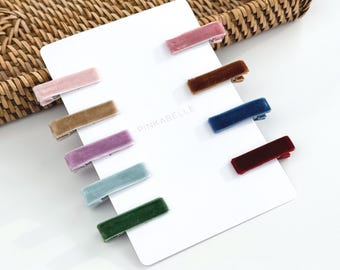 May include: A collection of velvet hair barrettes in various colors, including pink, brown, blue, and green, arranged on a white card. The barrettes are rectangular and have a shiny texture. The brand name "PINKABELLE" is printed on the card.