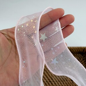 May include: A sheer pink ribbon with a delicate star pattern and a pink stitched edge. The ribbon is slightly translucent, allowing light to pass through, and is ideal for gift wrapping or crafting projects.