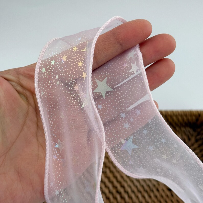 May include: A sheer pink ribbon with a delicate star pattern and a pink stitched edge. The ribbon is slightly translucent, allowing light to pass through, and is ideal for gift wrapping or crafting projects.