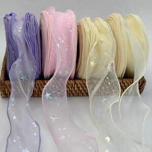 May include: Assortment of decorative ribbons in a woven basket. The ribbons are in shades of purple, pink, and cream, with a sheer overlay adorned with star-shaped glitter. The ribbons are ideal for crafts, gift wrapping, or adding a touch of elegance to any project.
