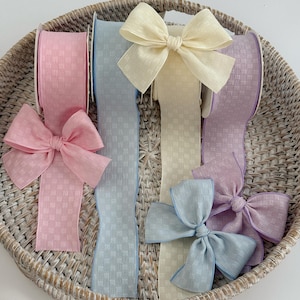 May include: A collection of decorative ribbons in various pastel colors, including pink, light blue, cream, and lavender. Each ribbon is textured with a checkered pattern and is tied into a bow. The ribbons are displayed in a woven basket.