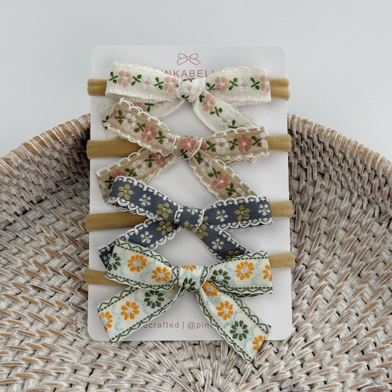 May include: A set of four floral patterned baby headbands. Each headband features a different color bow with a delicate floral design. The bows are attached to a soft, neutral-colored elastic band. The card reads "INKABEL" and "crafted | @pinkabel".