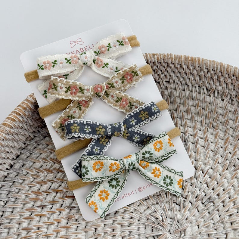 May include: Four floral patterned baby headbands on a white card. The headbands feature various colors, including white, beige, gray, and green, with floral embroidery and lace trim. The headbands are displayed on a woven basket.