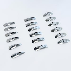 May include: A collection of silver-toned hair clips and barrettes arranged on a white surface. The clips are in various styles, including oval snap clips and slotted barrettes. The shiny metal reflects light, creating a clean and modern aesthetic.