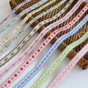 Könnte beinhalten: Eine Sammlung schmaler Stoffbänder mit verschiedenen Blumenmustern in Farben wie Rosa, Blau, Grün, Orange und Braun. Jedes Band hat einen gewellten Rand und ist diagonal angeordnet, wobei die verschiedenen gestickten Blumenmotive gut zu erkennen sind.