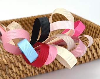 May include: A collection of colorful grosgrain ribbons in various shades of pink, red, blue, black, and cream, arranged in loops. The ribbons are displayed in a woven basket, creating a visually appealing display for crafting or gift wrapping.