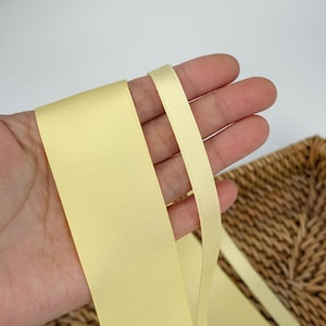 Könnte beinhalten: Hellgelbes Satinband in verschiedenen Breiten. Das Band wird in einer Hand gehalten, mit einem Weidenkorb im Hintergrund. Das Band hat eine glatte, glänzende Oberfläche, ideal für Bastelarbeiten, Geschenkverpackungen oder dekorative Zwecke.