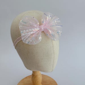 May include: A pink headband with a large, sparkly bow. The bow is made of tulle and has iridescent sequins.
